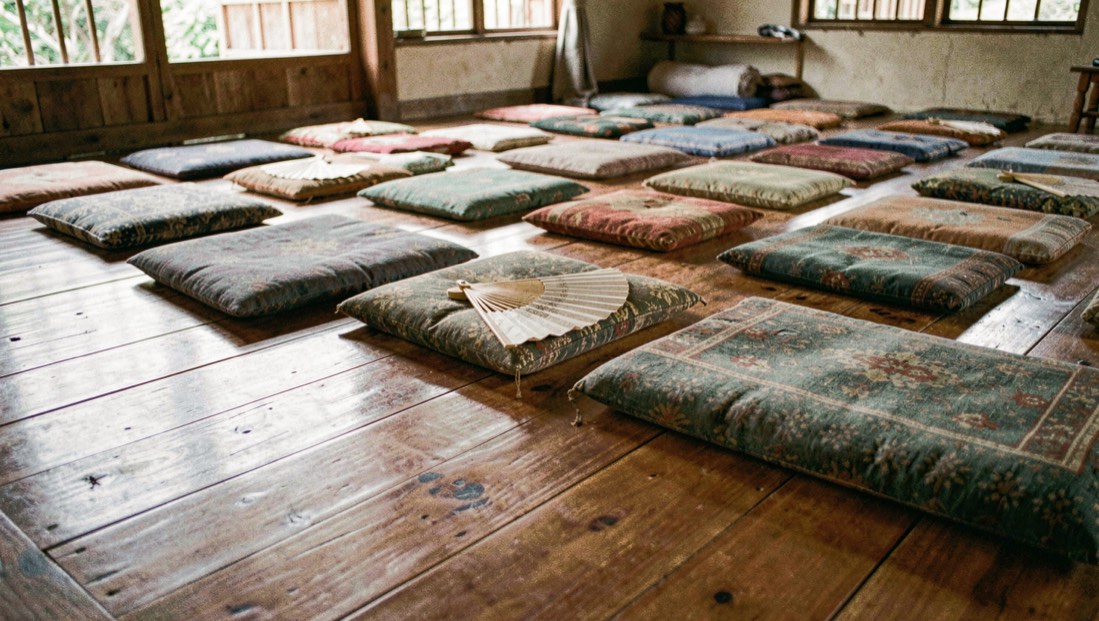 Cushions in tidy rows on the polished teak floor of a village meditation hall with a folded fan resting beside each seat in morning quiet